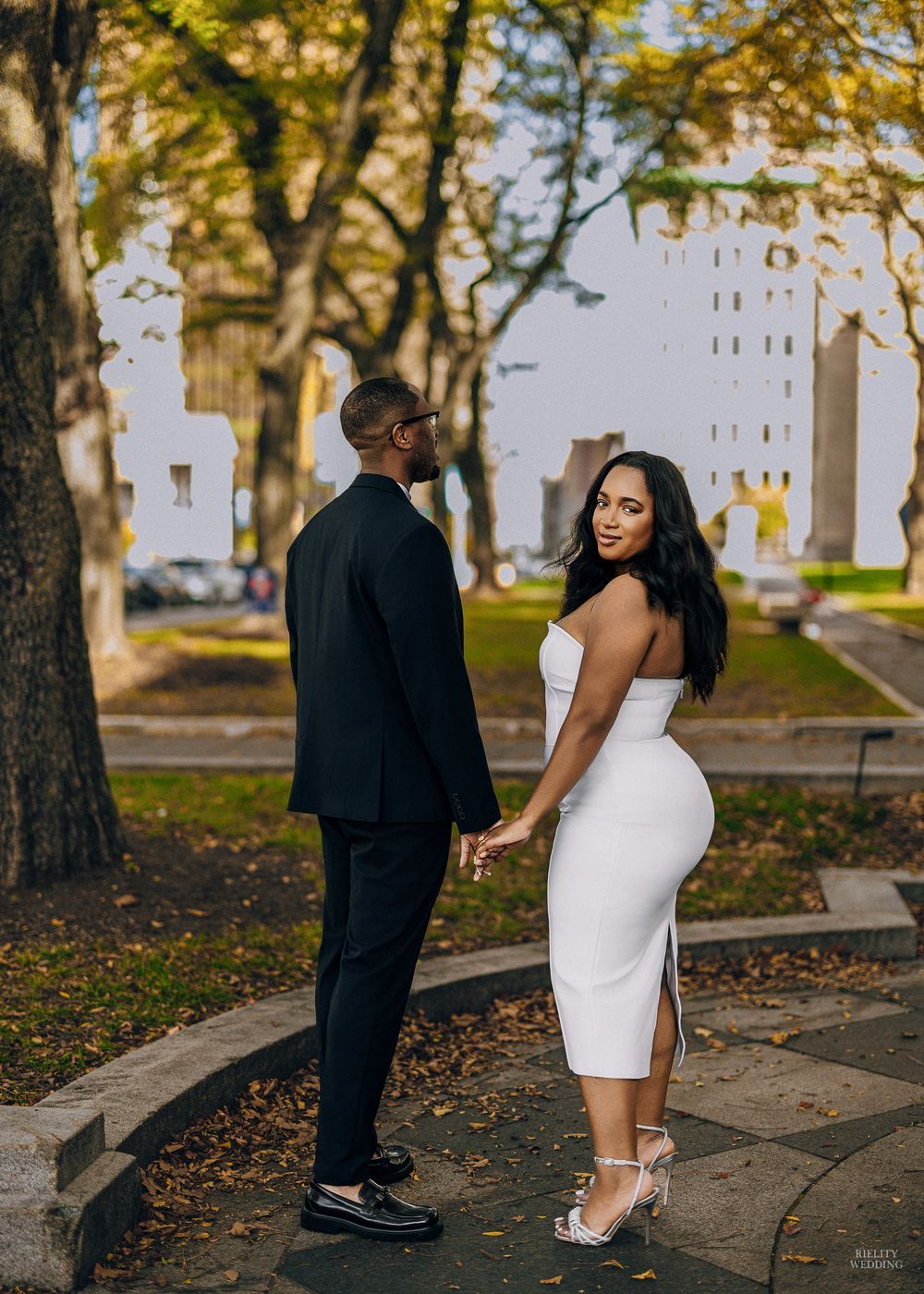 A couple poses playfully together on a tree-lined path during an autumn photoshoot.