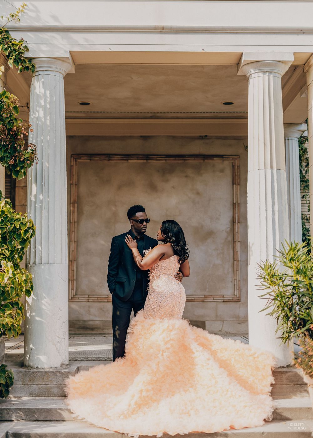 A couple in formal attire poses together under a columned portico with a flowing peach-colored gown creating a dramatic effect.