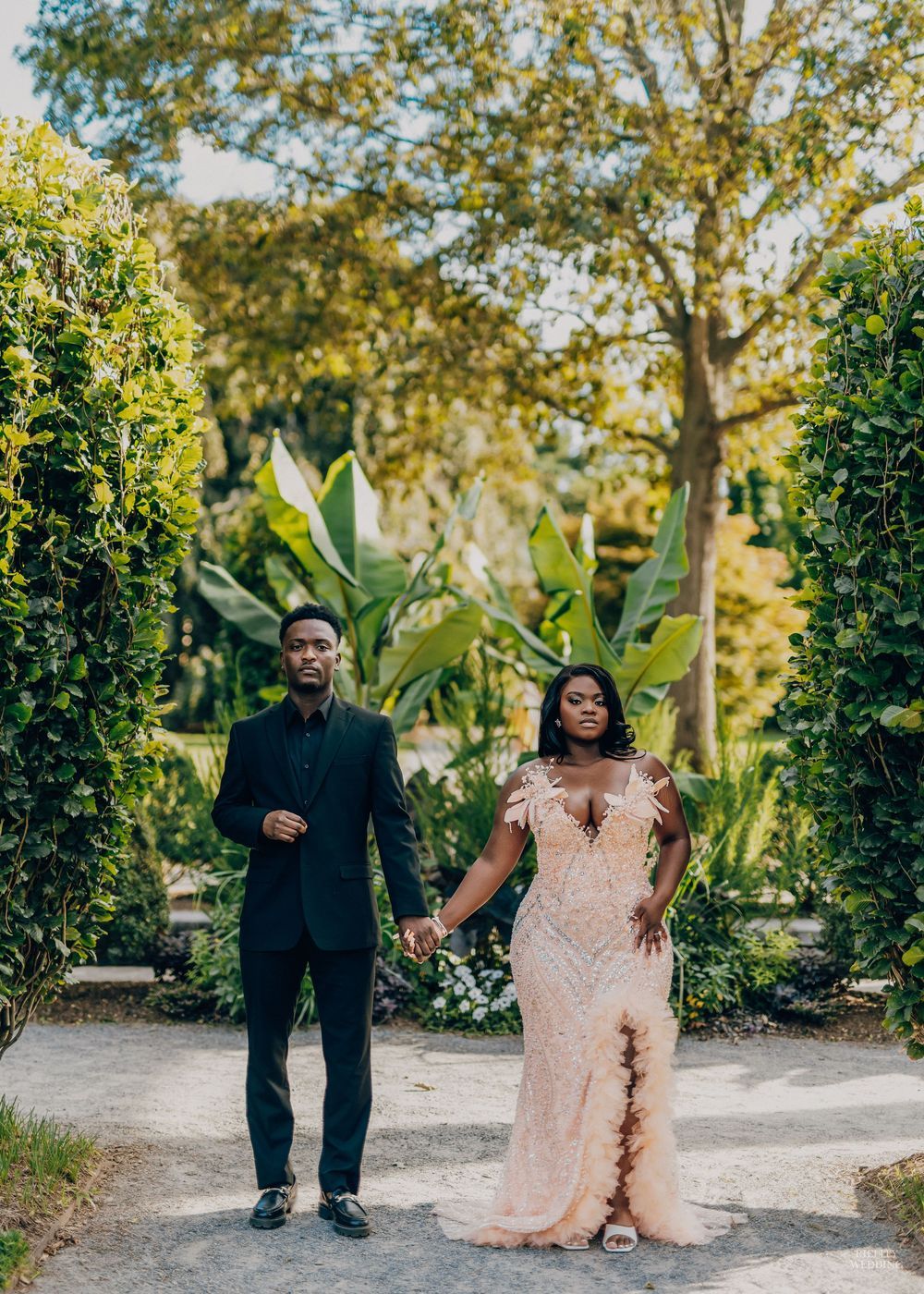 A couple poses separately in a lush garden setting wearing formal attire with pink gown and black suit.