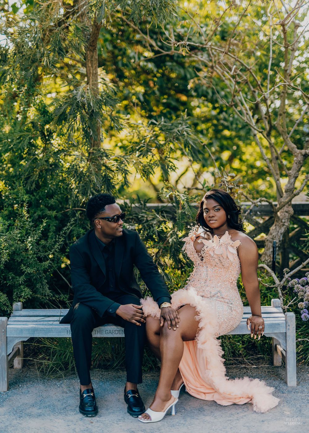 A couple sits together on a white bench in a garden setting wearing contrasting formal attire in black and peach.