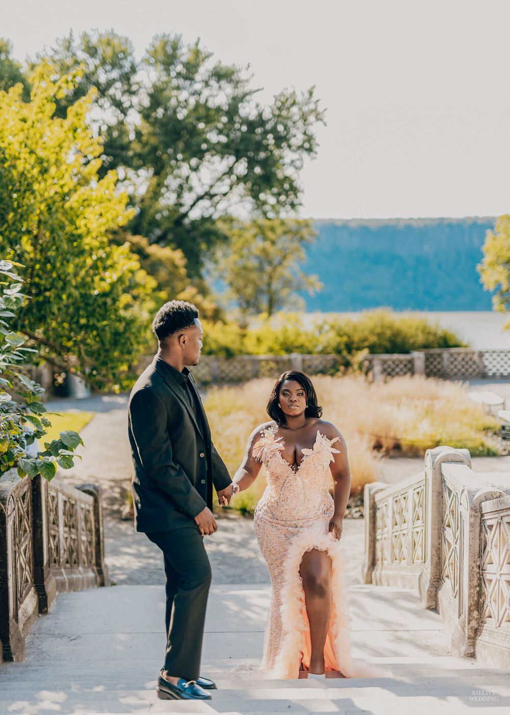 A couple shares a joyful moment on stone steps surrounded by yellow flowers and trees near a waterfront.