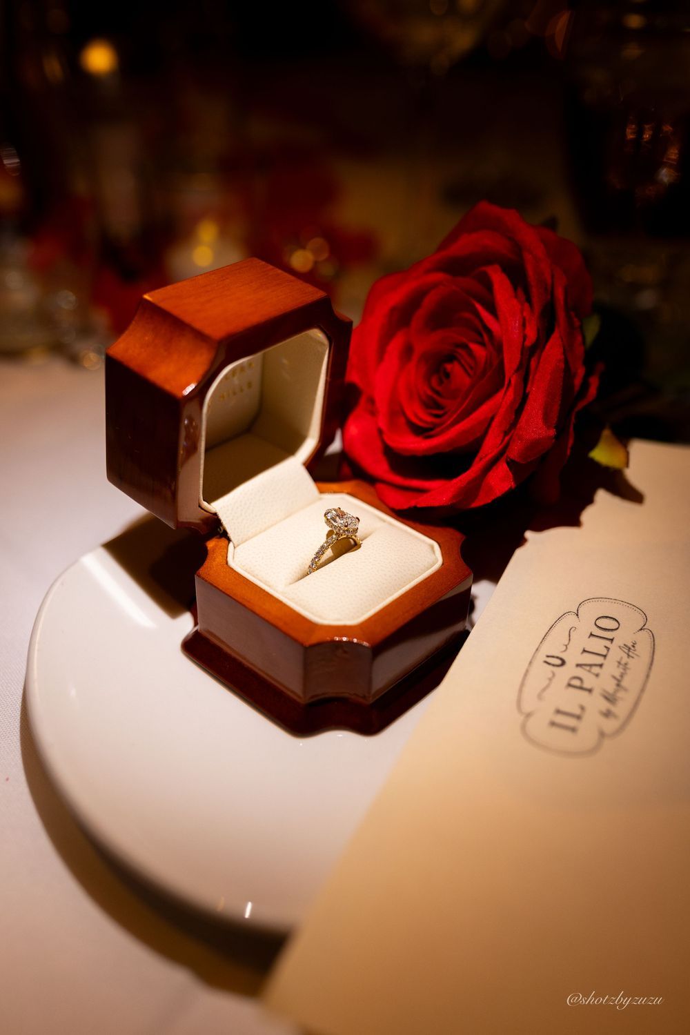 An engagement ring in a brown box next to a red rose on a white surface.