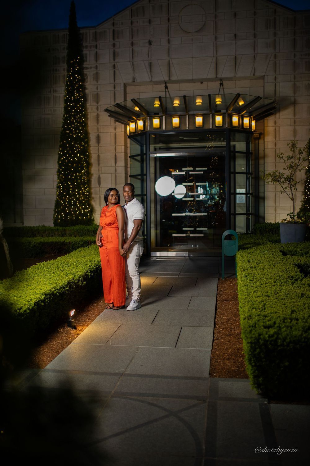 A couple stands on pathway with illuminated entrance and decorative trees at dusk.