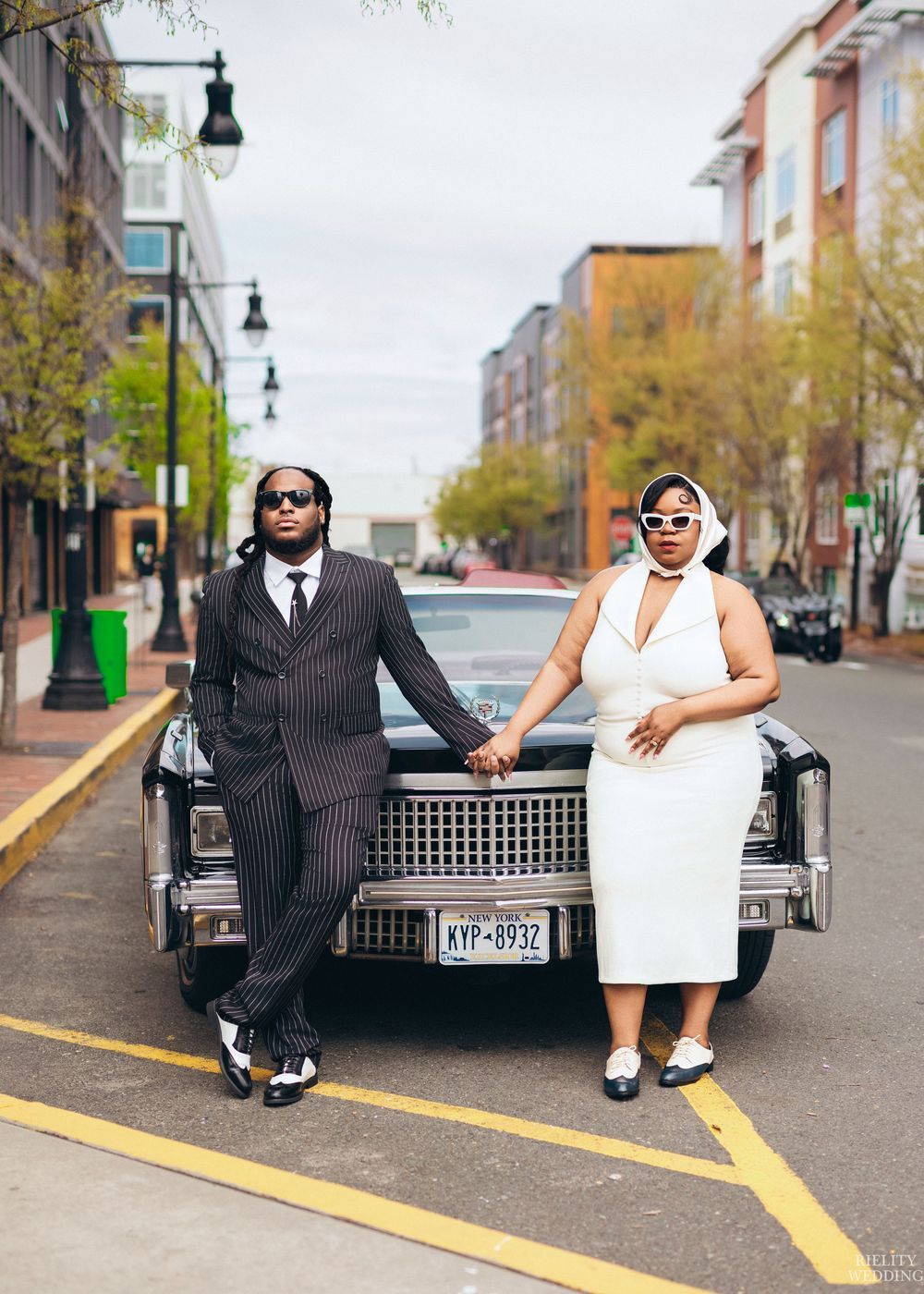 A stylish couple poses with a vintage car on a city street wearing black and white formal attire.