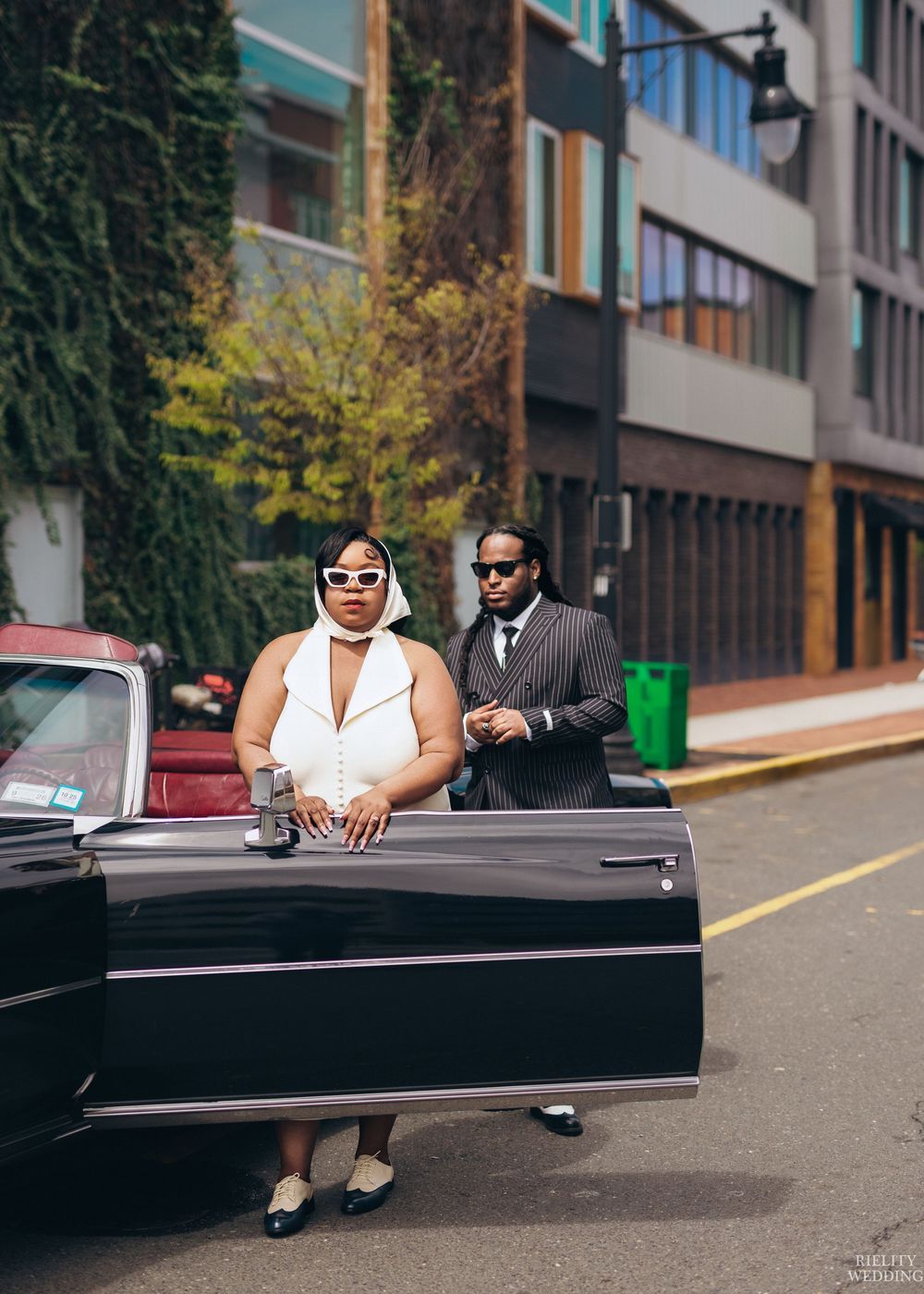 A couple in formal attire poses next to a sleek black vintage car on a city street.