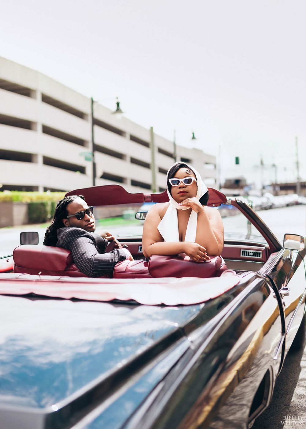 A person relaxes and poses in a vintage burgundy convertible car wearing white sunglasses.