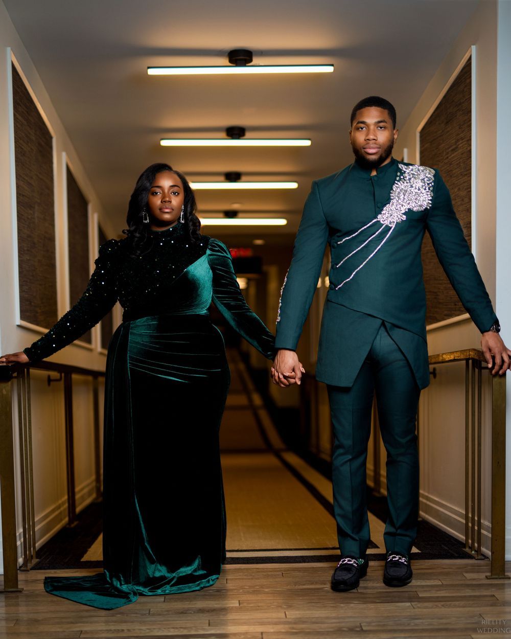 A couple in matching emerald green formal wear walks down a hallway hand in hand.