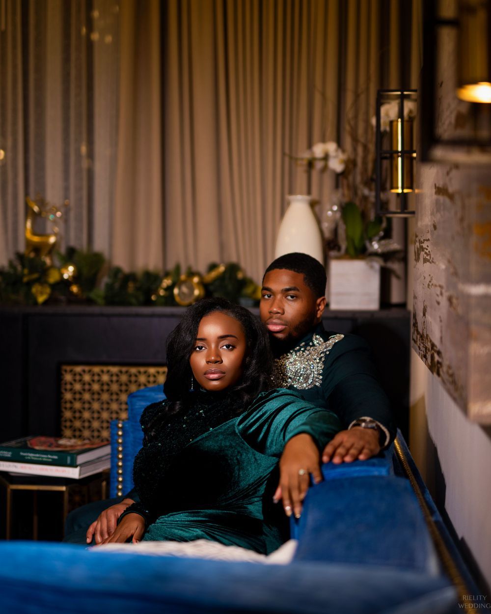 Couple wearing emerald green velvet outfits relaxing on blue couch with decorative lighting and garland in background.