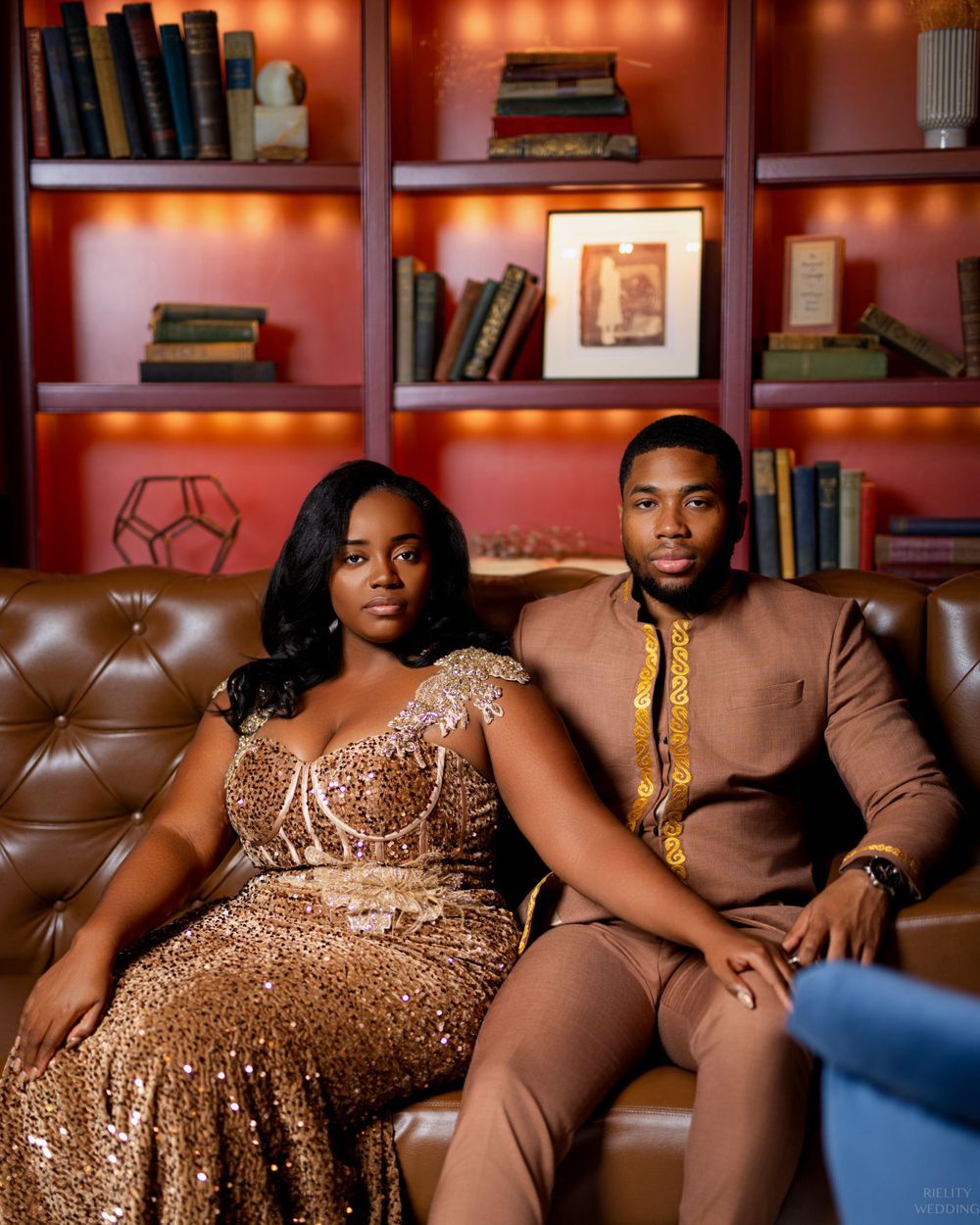 A couple poses elegantly in formal wear against a luxurious red bookshelf backdrop.