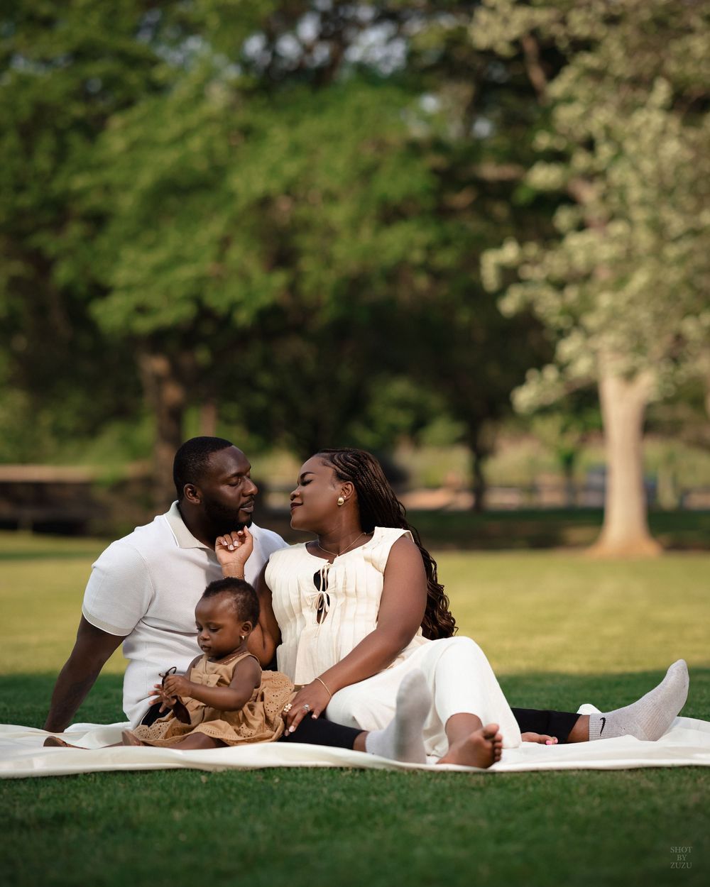 A family lies together on a blanket in a sunny park setting, sharing intimate moments and laughter.