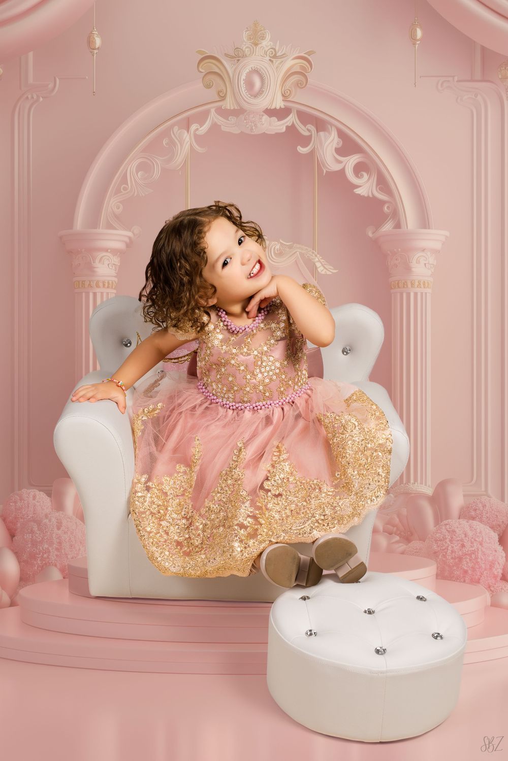 A child in a sparkly pink and gold dress poses playfully on a white chair against a pink backdrop.