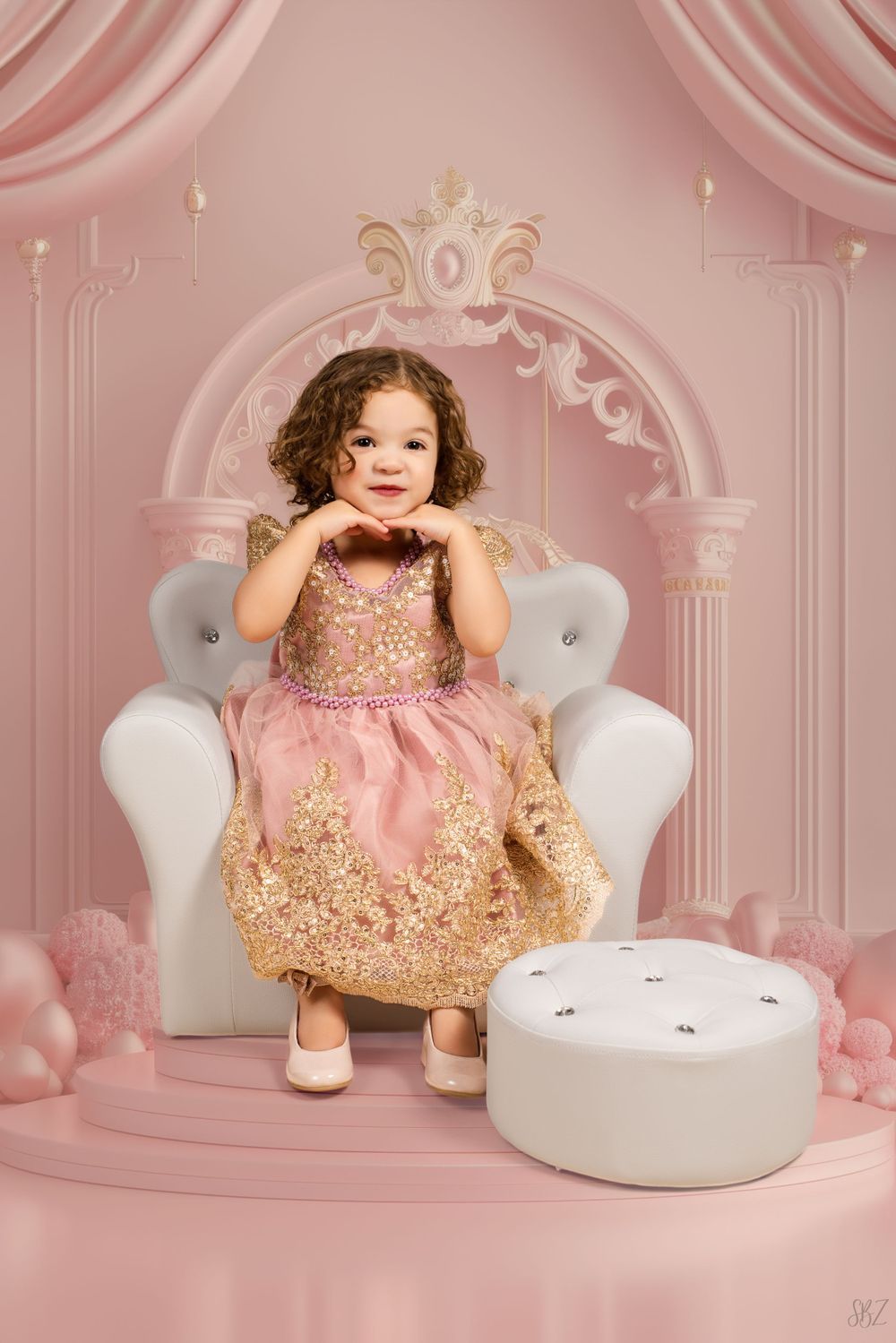 A child poses sweetly in a pink and gold dress on a white throne chair with pink decorative backdrop.