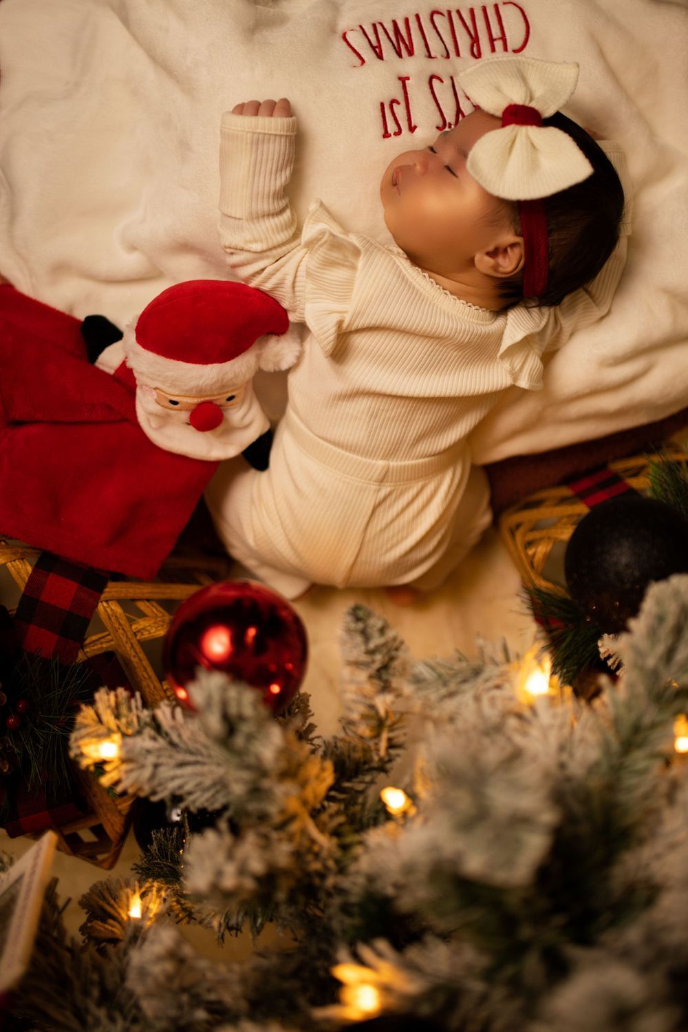 Baby sleeping on white Christmas blanket next to Santa plush toy with decorated tree and lights in background.