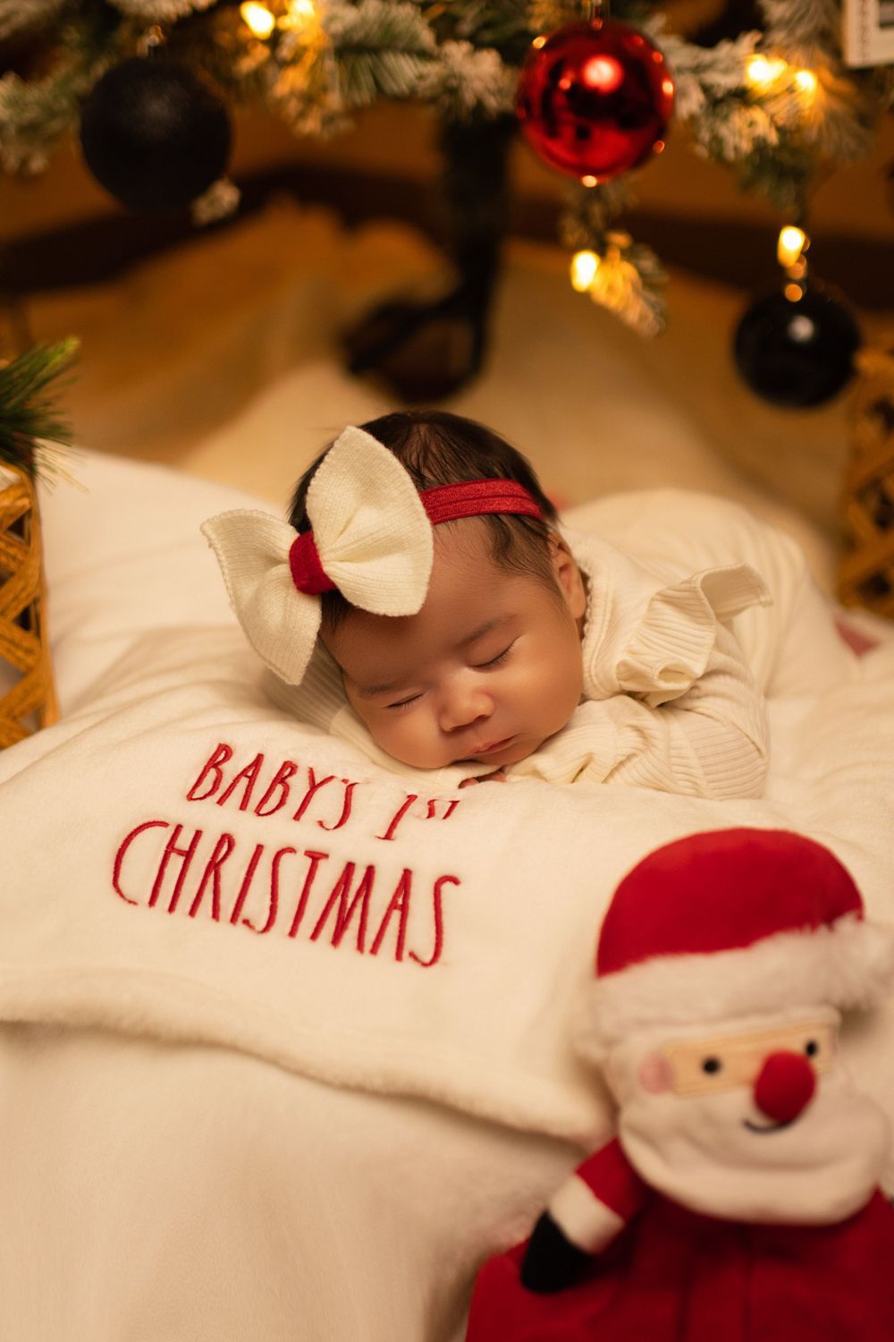 Peaceful baby napping on festive white pillow next to Santa doll with Christmas decorations overhead.