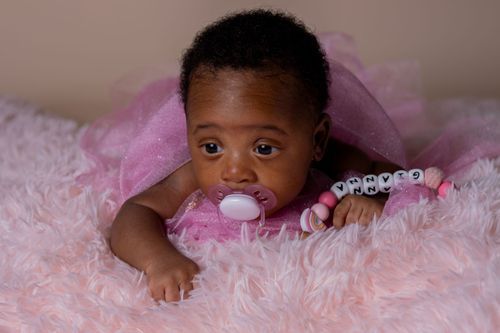 Baby with pink pacifier and beaded necklace lying on pink furry blanket.