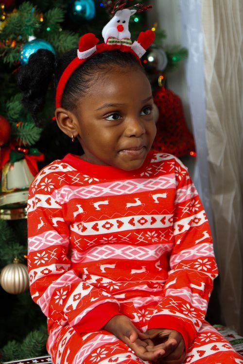 Child wearing festive red and white patterned sweater and reindeer headband poses by Christmas tree.