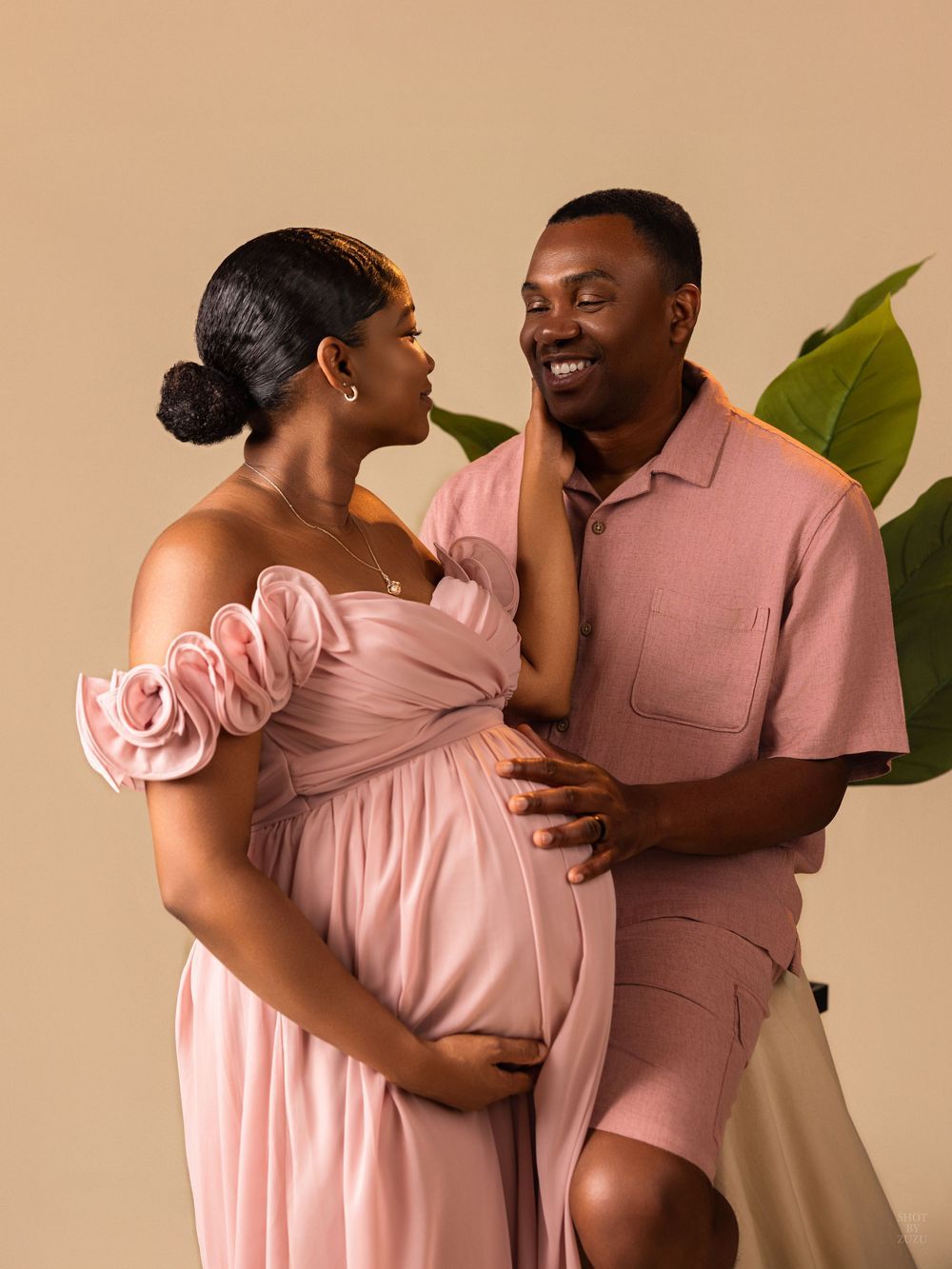 A couple in matching pink outfits shares intimate moments in a romantic photoshoot against a neutral background.