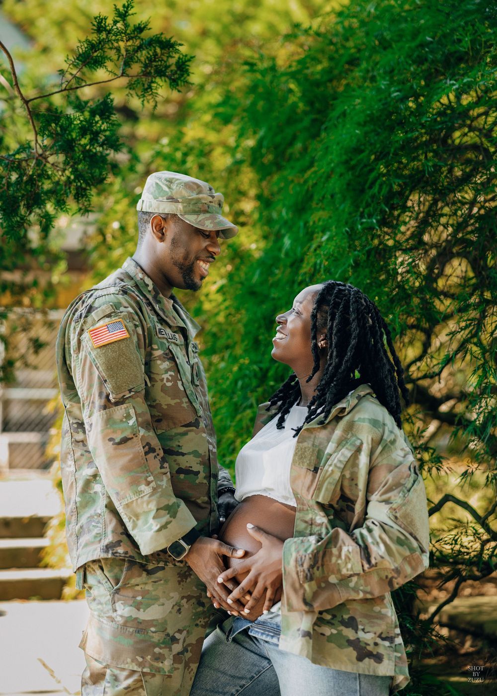 Military couple in camouflage uniforms share tender moments in a lush green outdoor setting.