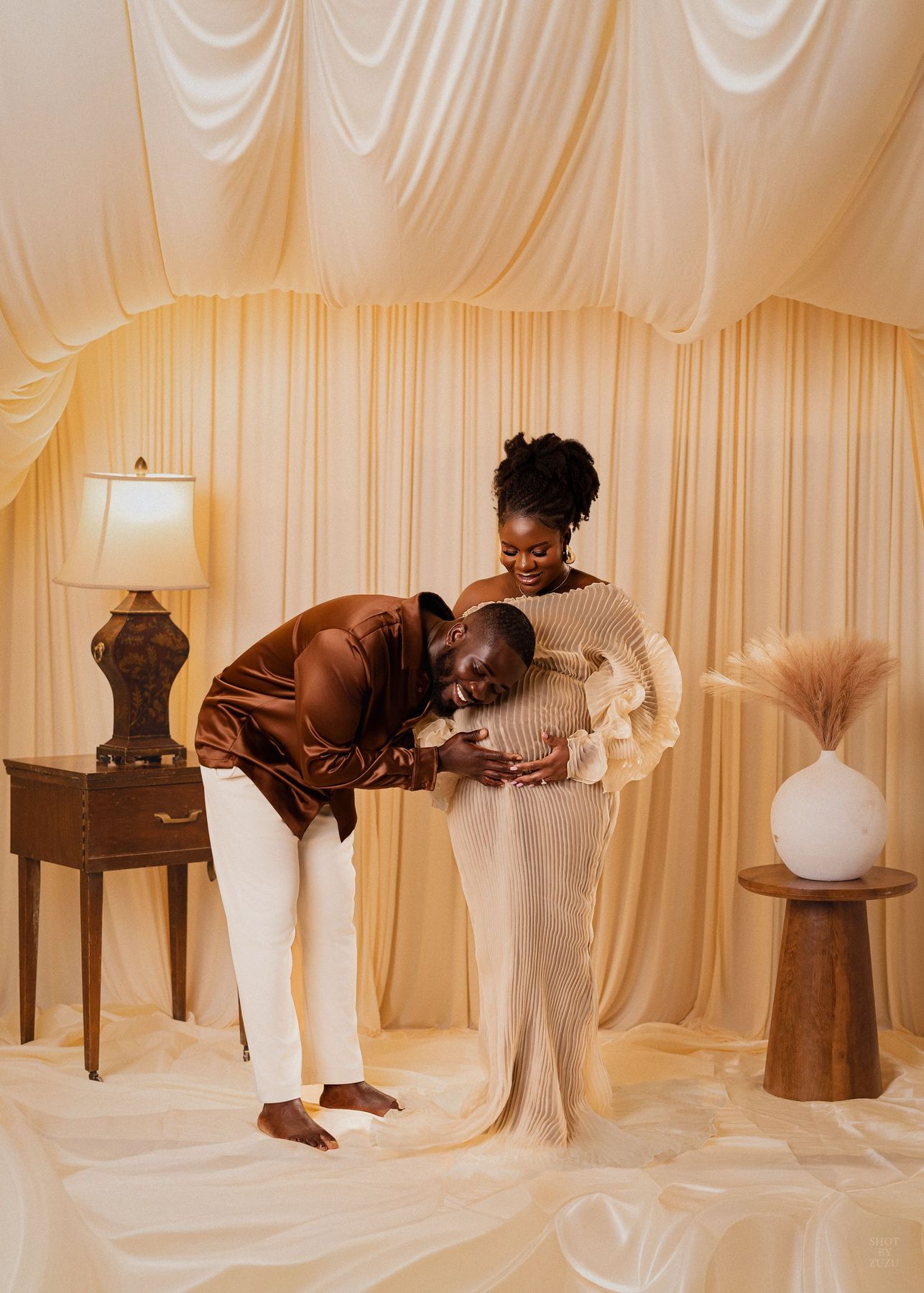 A couple shares a tender maternity moment, man kissing baby bump, in an elegant cream-draped studio setting.