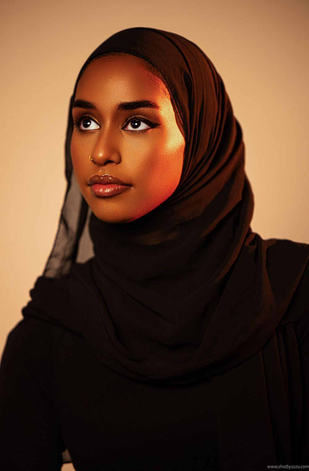 Portrait of woman wearing black hijab against warm-toned background with dramatic lighting.