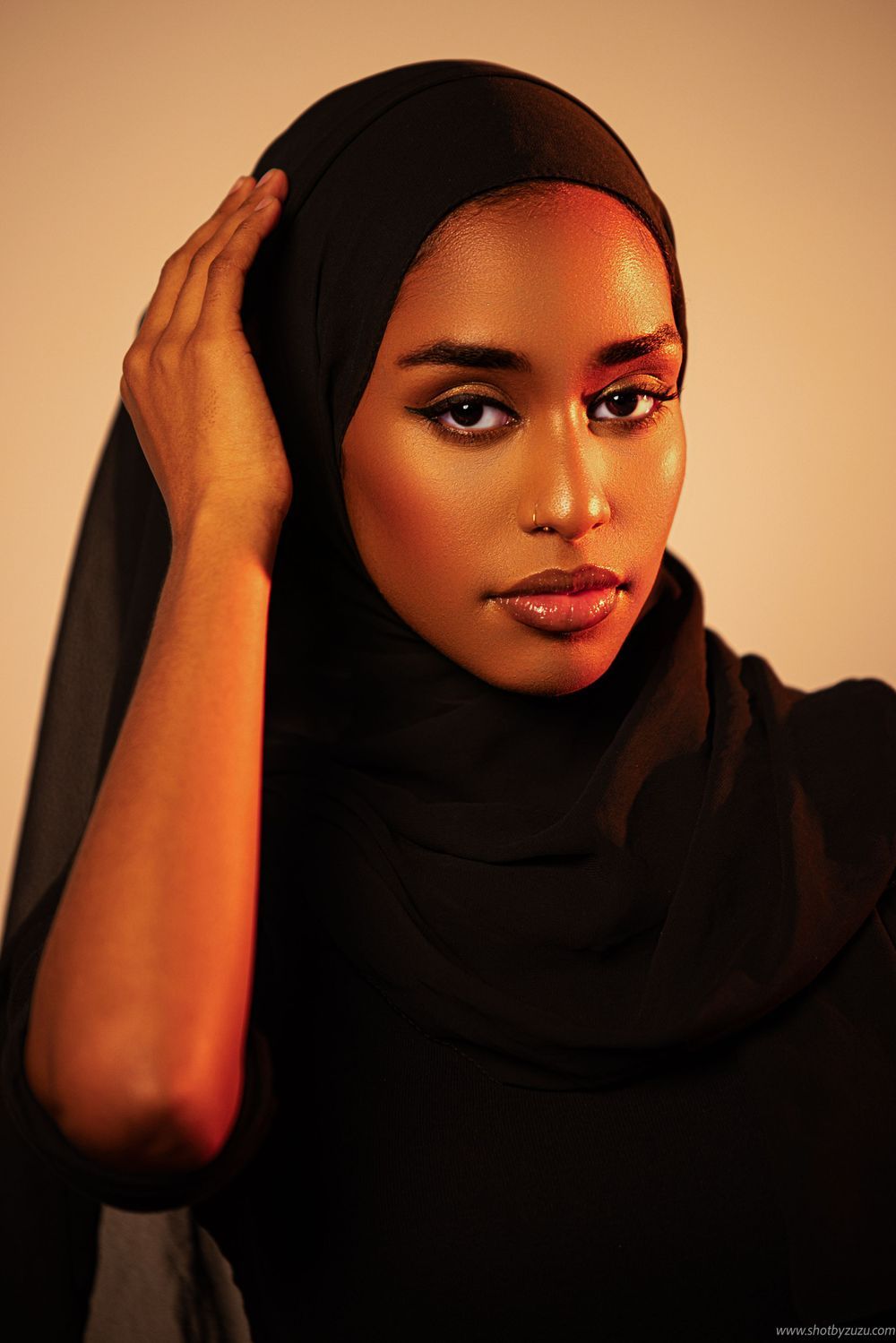 Glamorous portrait of woman in black hijab with soft lighting and warm background tones.