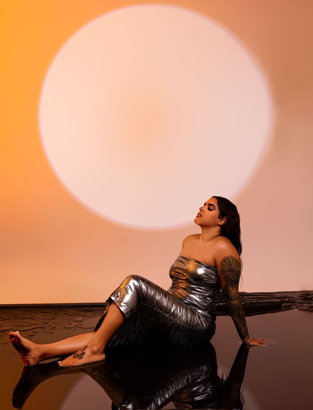 Fashion portrait with silver metallic dress against large orange circular backdrop.