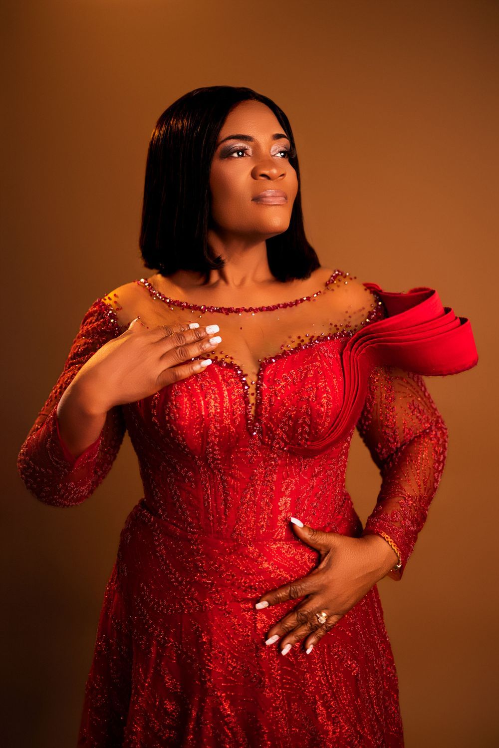 A person poses in a glamorous red beaded gown with sheer sleeves against a warm brown backdrop.