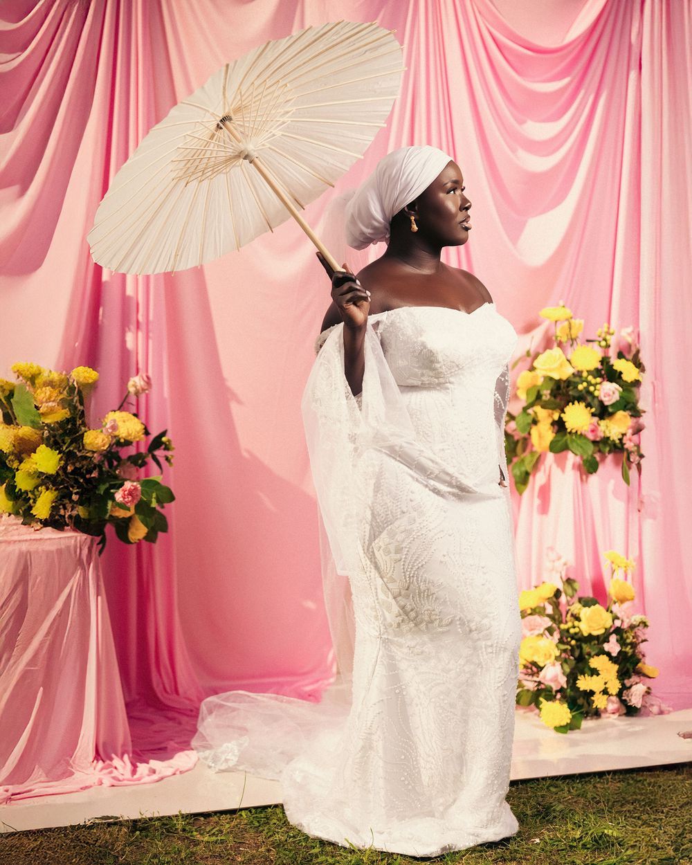 Bride in white lace wedding gown poses with parasol against pink draped backdrop with yellow floral arrangements.