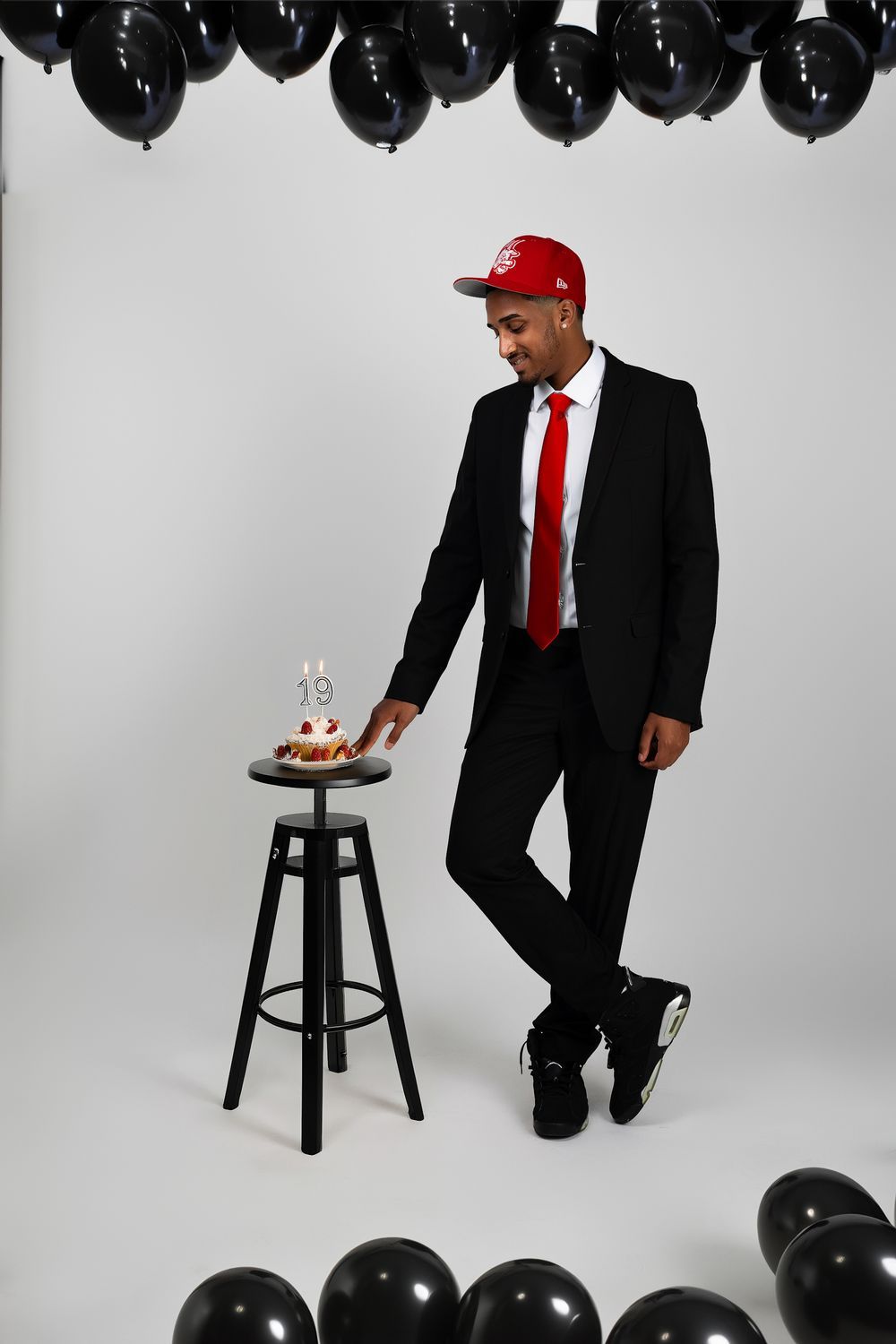 Studio portrait with black balloons featuring formal suit, red cap and tie with birthday setup.