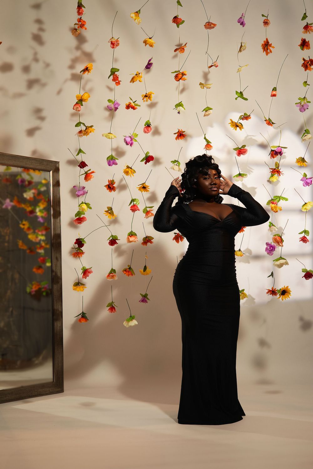 Woman in elegant black dress poses against wall decorated with colorful hanging flowers.