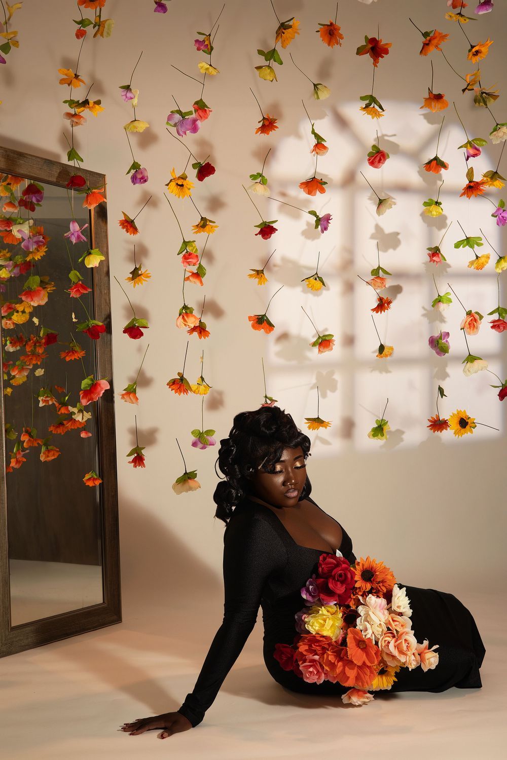 Woman in long black dress reclines on floor with vibrant floral bouquet against flower wall backdrop.