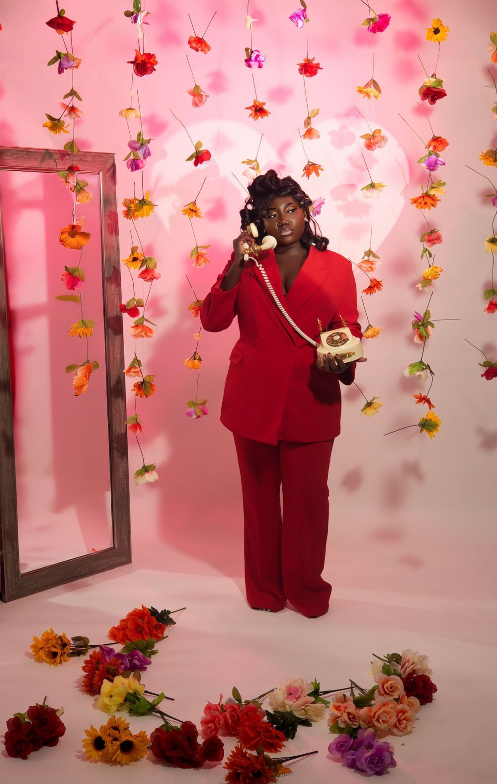 A bright red powersuit ensemble photographed against a pink backdrop with hanging floral decorations and a vintage telephone.