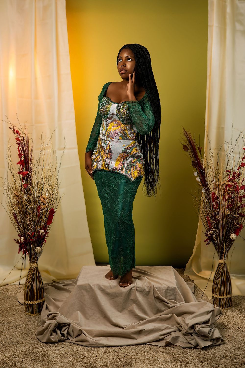 Model in emerald green gown with floral bodice poses against yellow backdrop with dried floral arrangements.