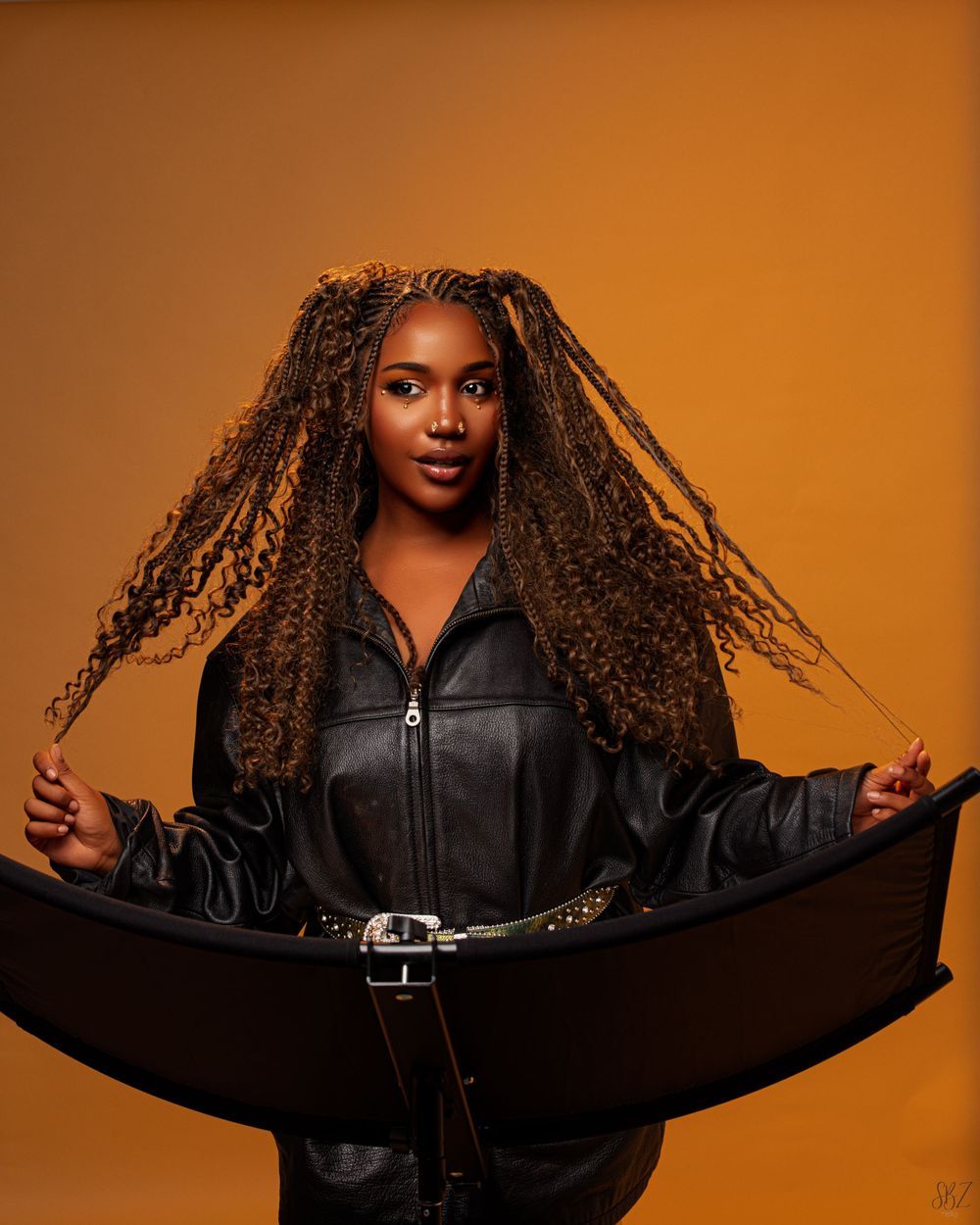 Fashion model in black leather jacket showcasing long braided hairstyle against orange backdrop.