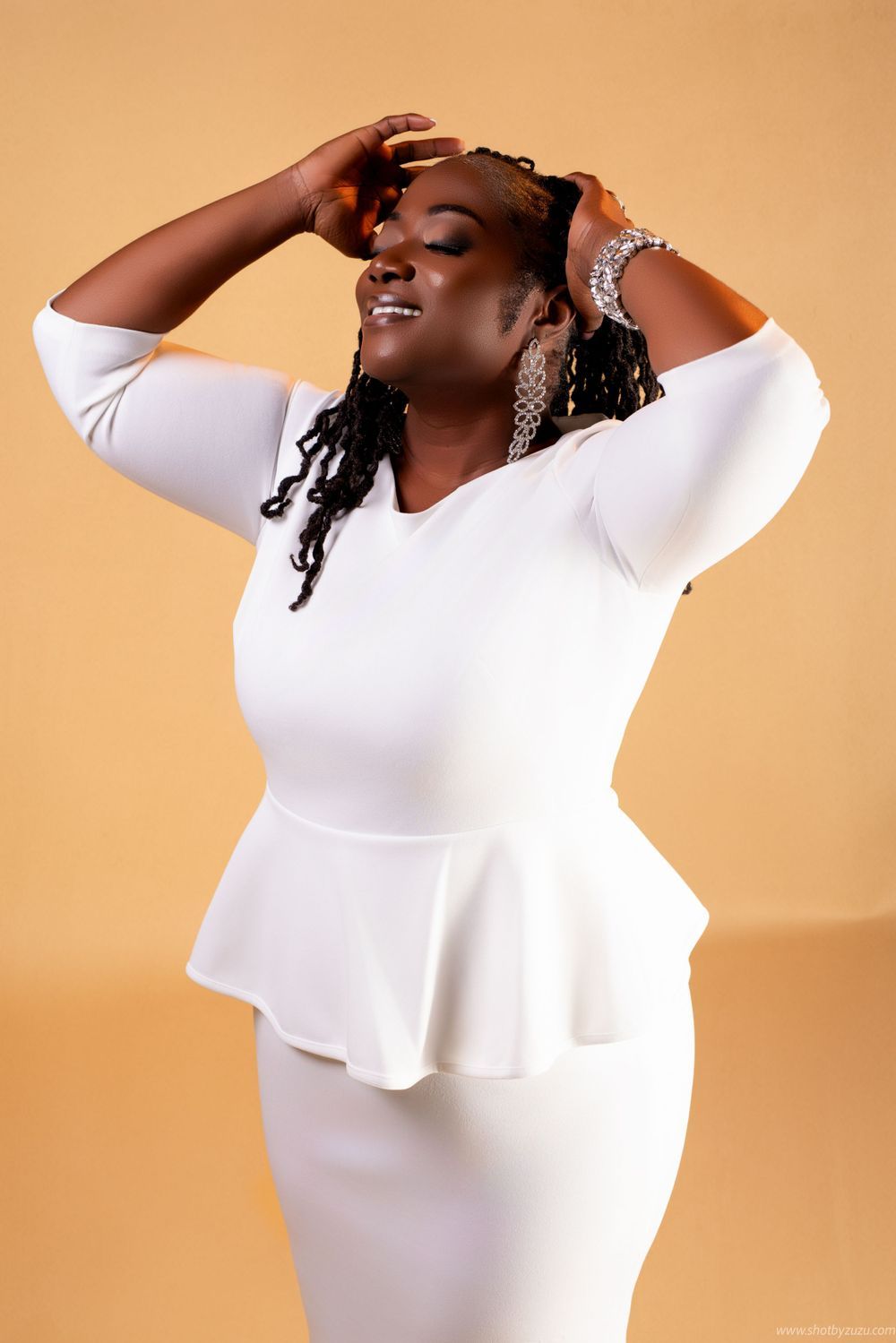 Joyful pose in white peplum dress with sparkling jewelry against neutral background.
