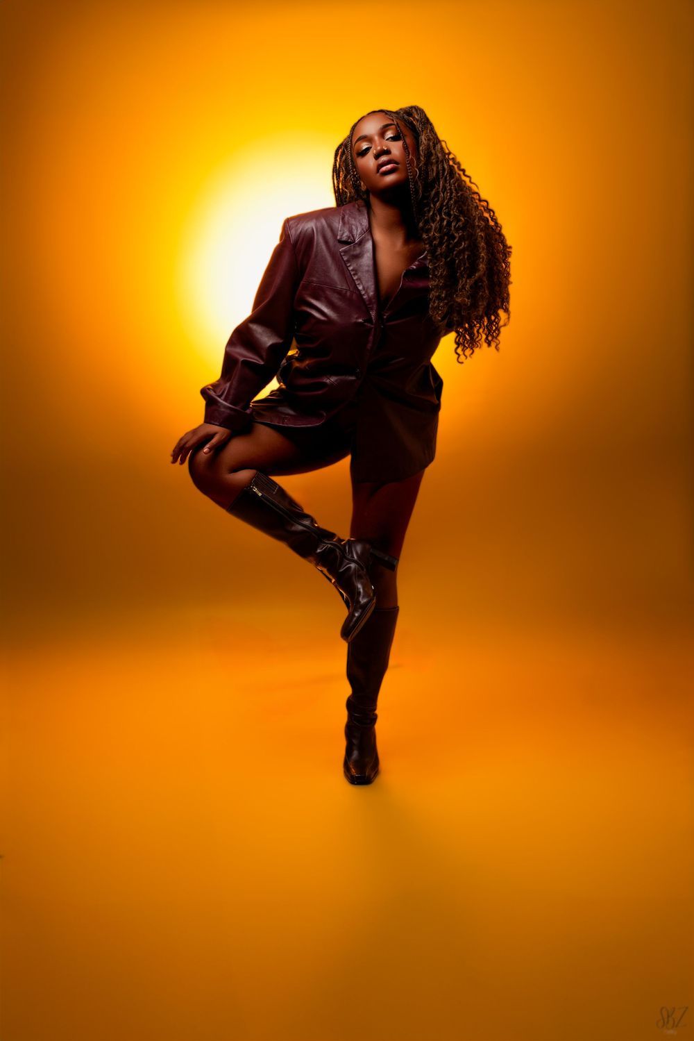 Dynamic pose in burgundy leather and boots against vibrant orange studio lighting.