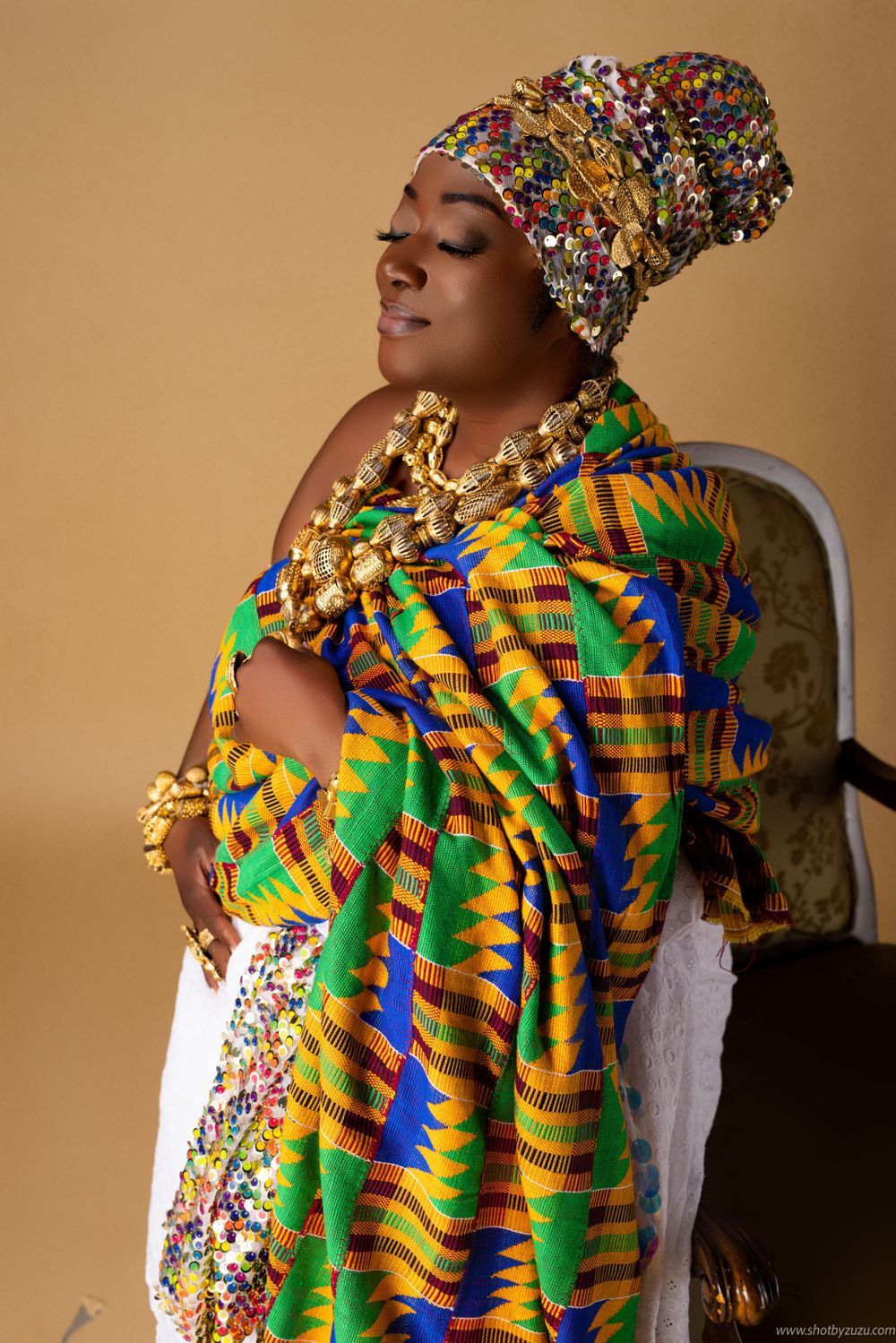 Woman in vibrant African print dress and headwrap poses against beige background.