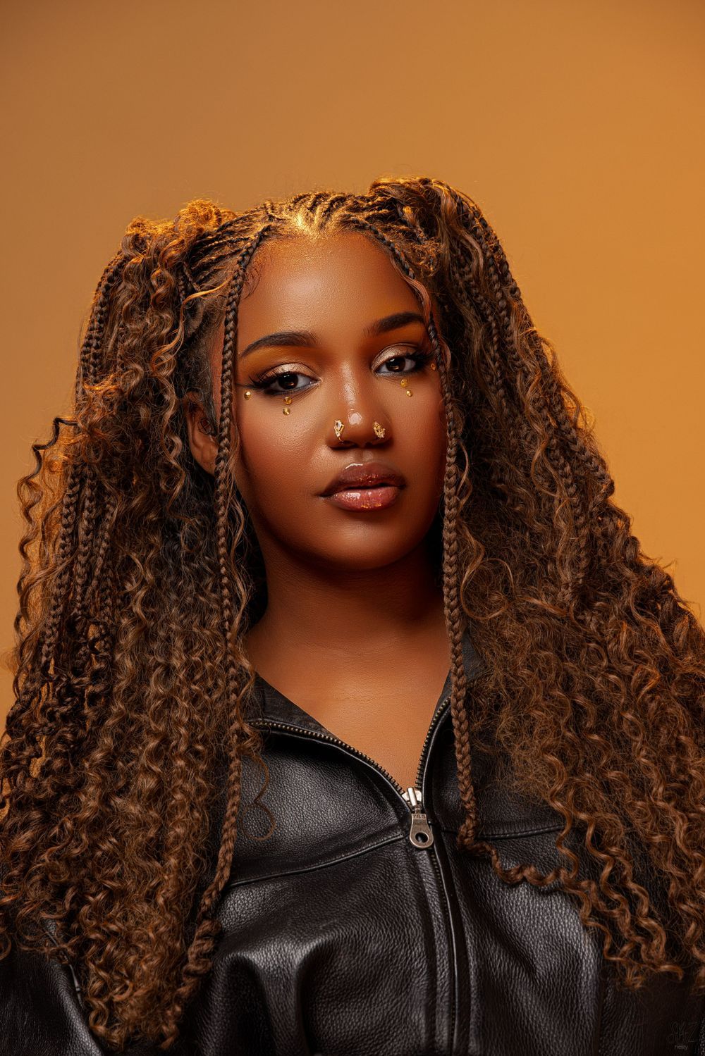 Editorial style portrait with textured braids and leather jacket against warm orange background.