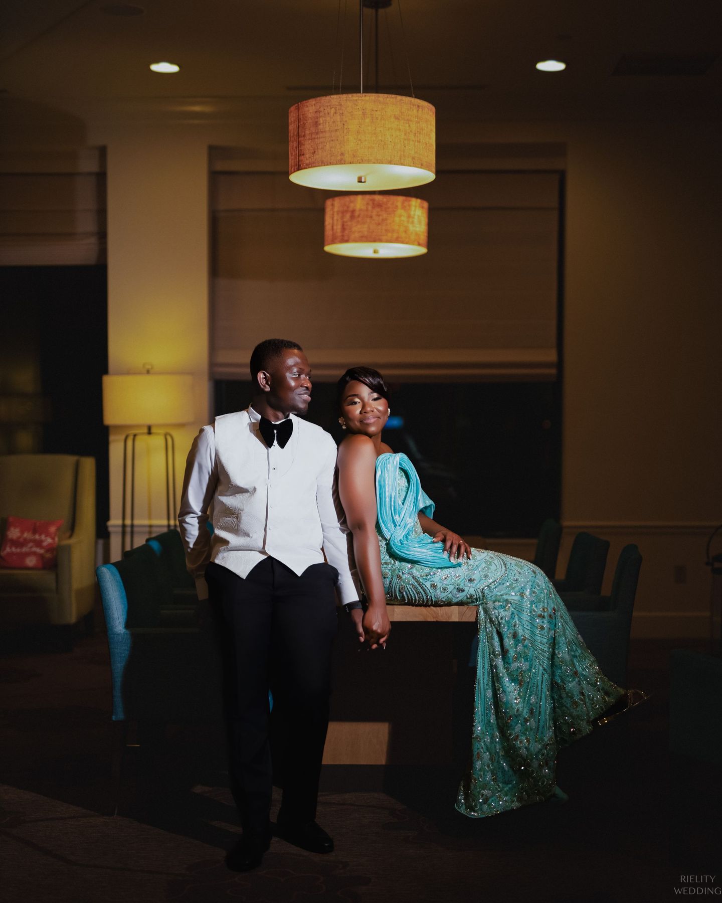 Couple poses together under pendant lights with groom in tuxedo and companion in teal gown.
