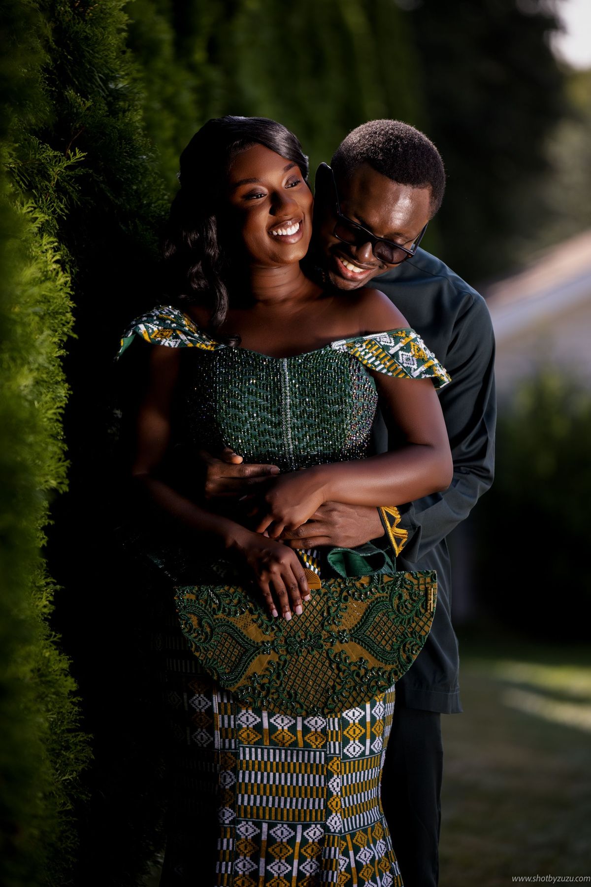 Couple in African dress embracing against green hedge background.